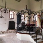 Historický interiér synagógy s kovanou bimou, ozdobnými lustrami a oblúkovými oknami s hebrejskými nápismi. Na stenách sú staré náboženské tabule a prepracované dekoratívne prvky.